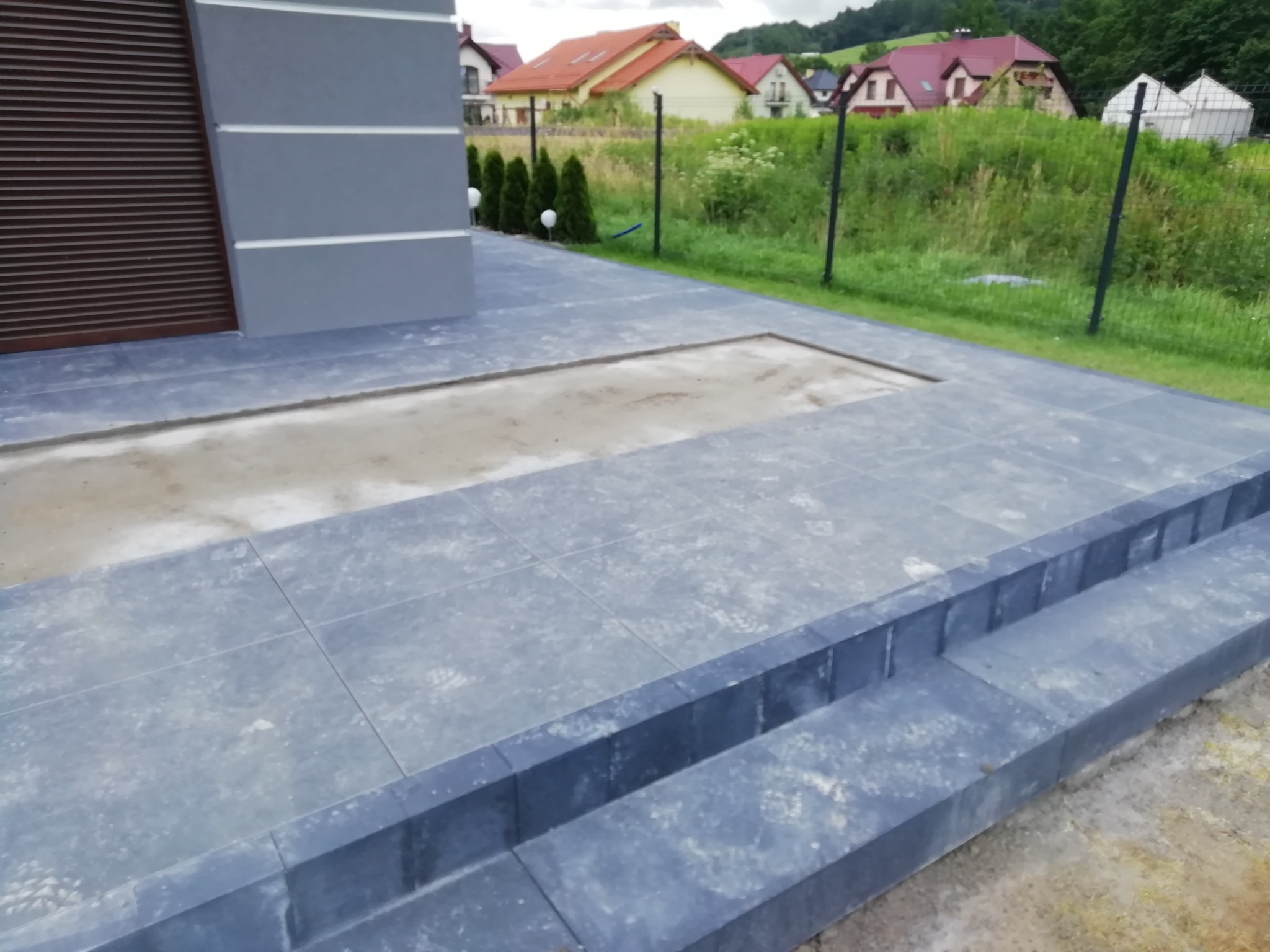 Szare płytki tarasowe ułożone na zewnątrz domu, z widocznym fragmentem przygotowanego podłoża w miejscu przyszłego trawnika lub rabaty.