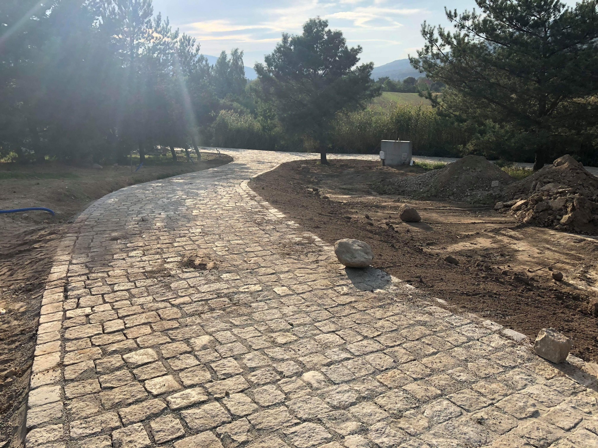 Zakrzywiona ścieżka z jasnej kostki, otoczona ziemią i zielenią, z widokiem na wzgórza w oddali, w słoneczny dzień.