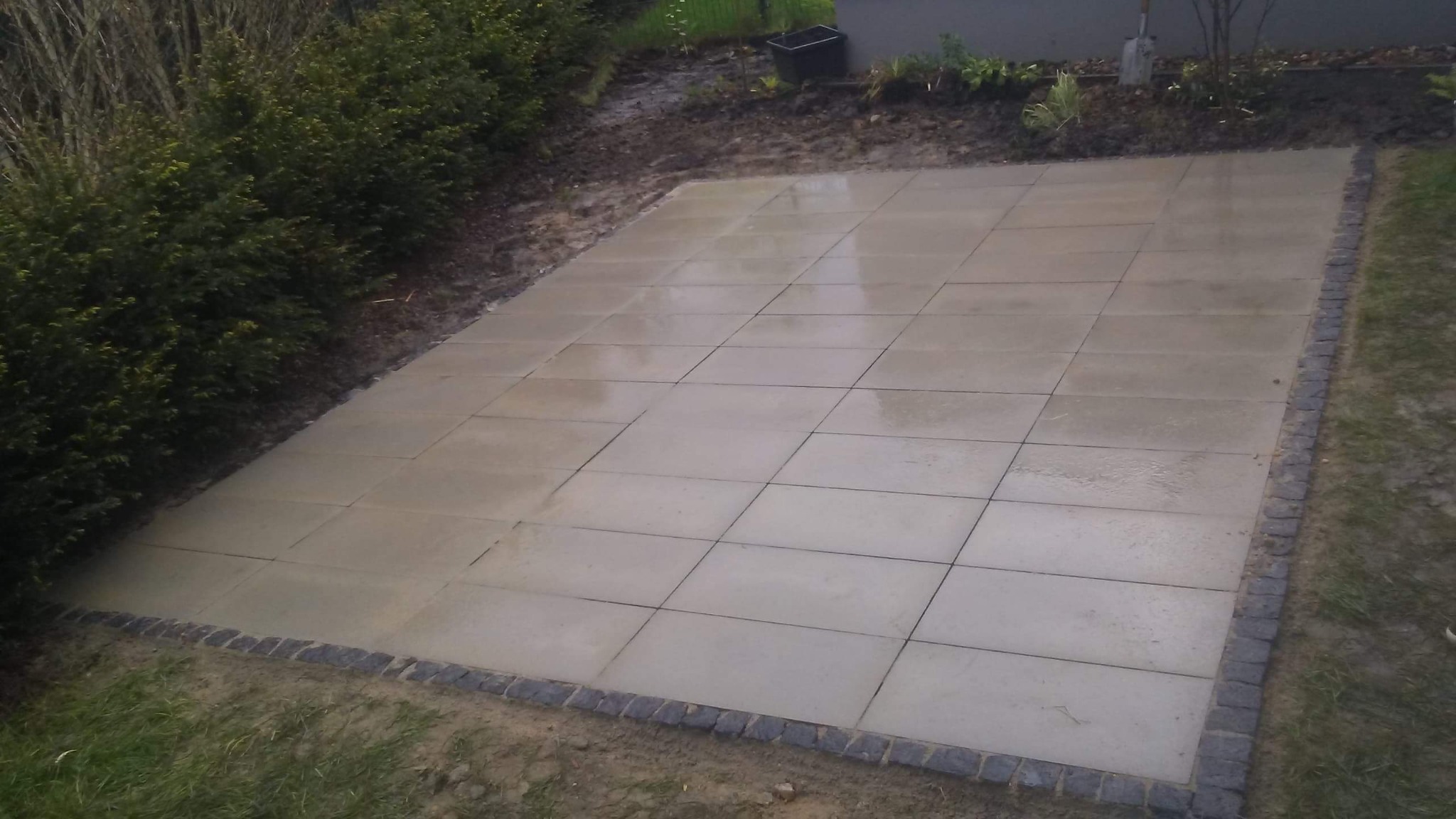 Nowo ułożony, prostokątny taras z jasnych płyt, obramowany ciemną kostką granitową. Tło stanowi trawa i krzewy.