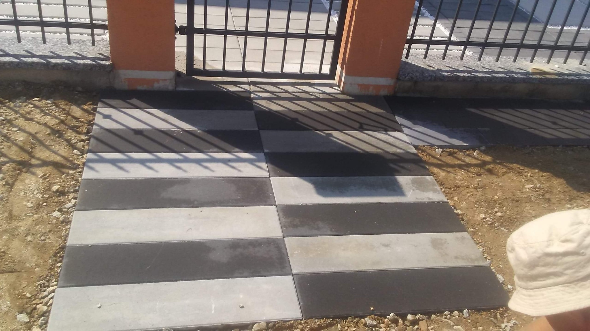 Ścieżka z szaro-czarnej kostki brukowej prowadząca do metalowej bramy z pomarańczowymi słupkami, widoczny fragment kapelusza w prawym dolnym rogu.