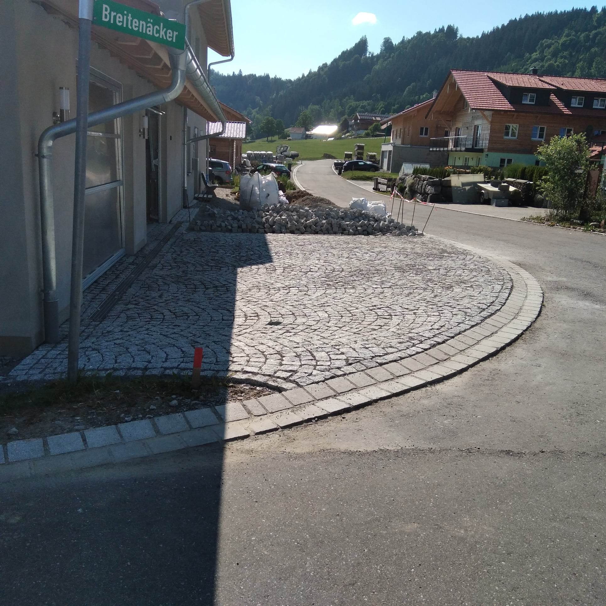 Układanie okrągłej powierzchni z kostki granitowej w formie wachlarza, z widocznymi workami z materiałem i zielonym znakiem ulicznym 'Breitenäcker' w tle.