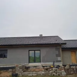 Dom jednorodzinny w trakcie budowy, z widocznym nowym dachem z ciemnej dachówki ceramicznej, rynny zamontowane, ściany zewnętrzne w stanie surowym, teren wokół domu z gruzem i materiałami budowlanymi.