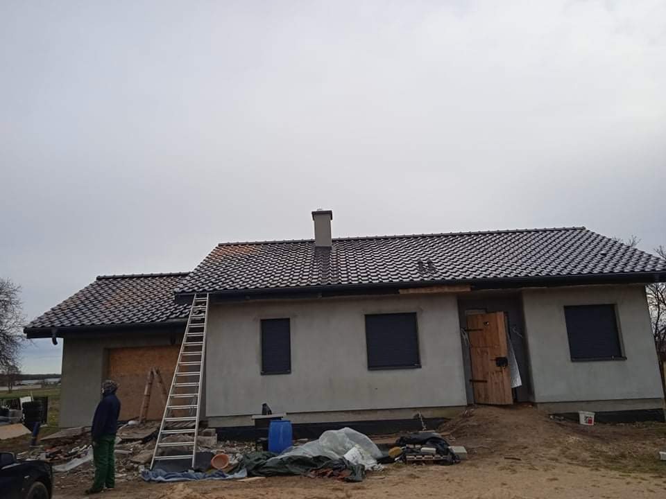 Dom jednorodzinny z nowym, ciemnoszarym dachem z dachówki ceramicznej, widoczna drabina oparta o ścianę i pracownik w oddali, teren wokół domu w trakcie prac wykończeniowych.