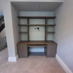 Wnęka z p&oacute;łkami i biurkiem z drewna w odcieniach szarości, widoczne gniazdko elektryczne bez osprzętu, marmurowa podłoga, białe listwy przypodłogowe i sufit z kratką wentylacyjną.