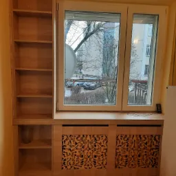 Drewniana zabudowa okna z ażurowym panelem grzejnika i regałem z półkami, widok na osiedle przez okno.