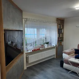 Wnętrze salonu z szarą podłogą, meblami na wymiar z szarymi frontami, półką na książki i oknem z firanką. W oknie widoczne zdjęcia i ozdoby.