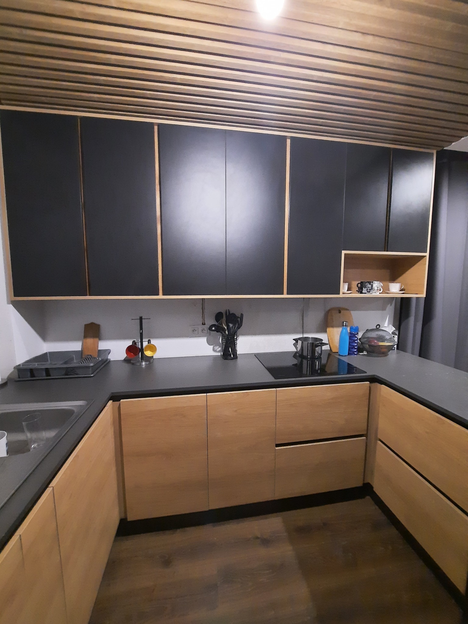 Nowoczesna kuchnia w Sędzinach z czarnymi szafkami górnymi i drewnianymi dolnymi, szary blat, drewniany sufit lamelowy i indukcyjna płyta grzewcza.