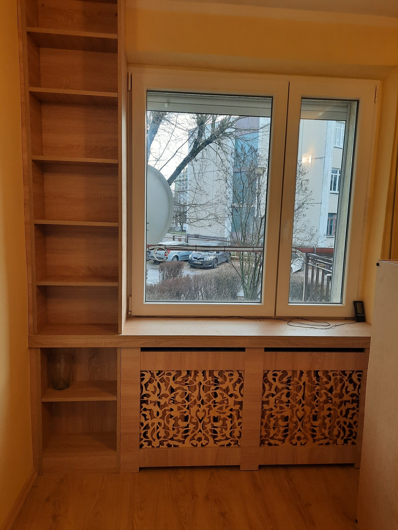 Drewniana zabudowa okna z ażurowym panelem grzejnika i regałem z półkami, widok na osiedle przez okno.