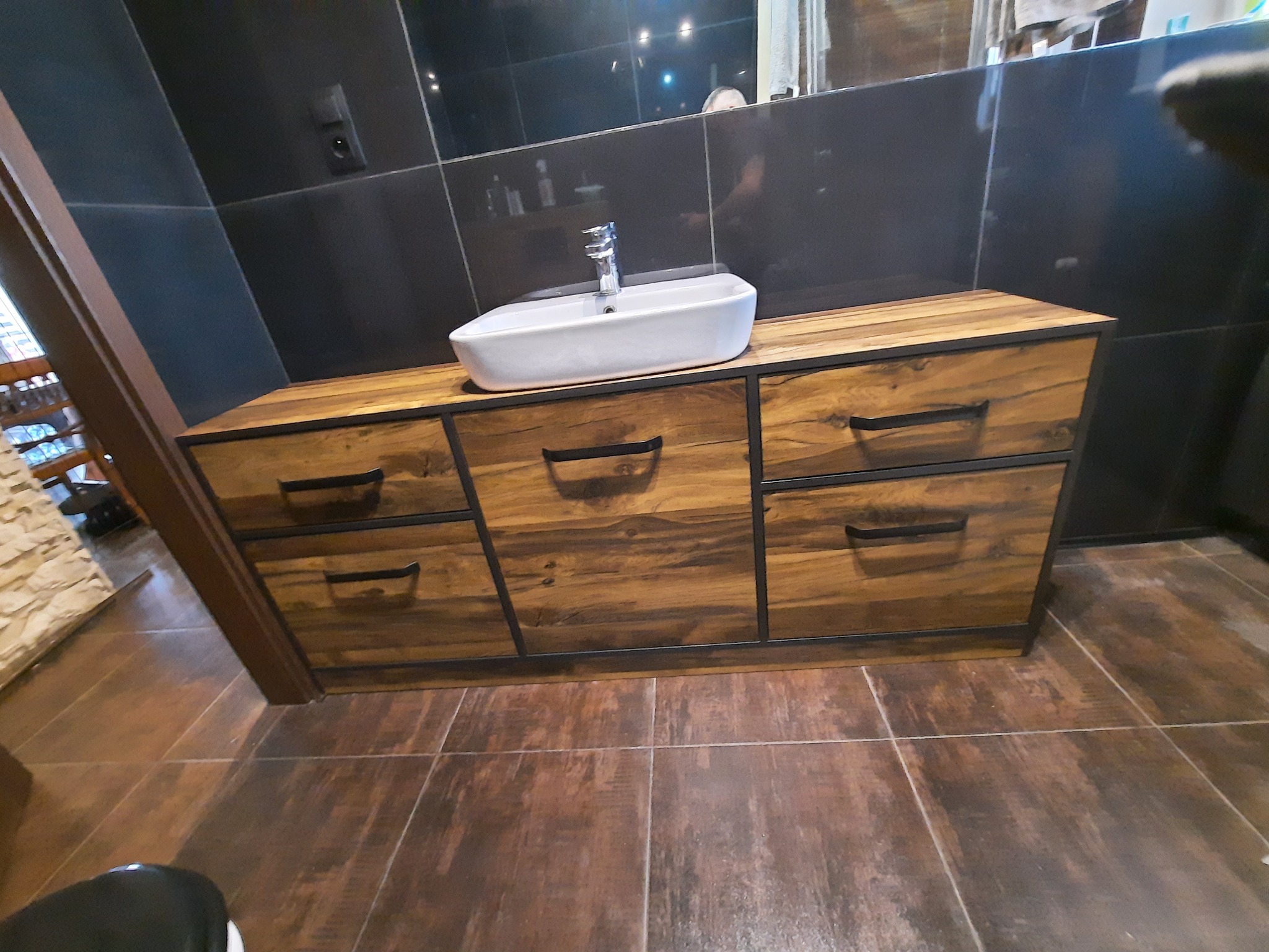 Szafka łazienkowa z drewnianymi frontami i czarnymi uchwytami, na niej biała umywalka nablatowa z chromowaną baterią, tło z ciemnych płytek.