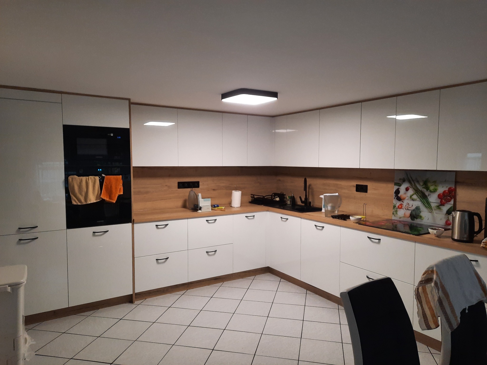 Nowoczesna kuchnia z białymi szafkami i drewnianym blatem, czarny piekarnik w zabudowie, płytki podłogowe w kratkę, oświetlenie LED w suficie, widoczne akcesoria kuchenne.