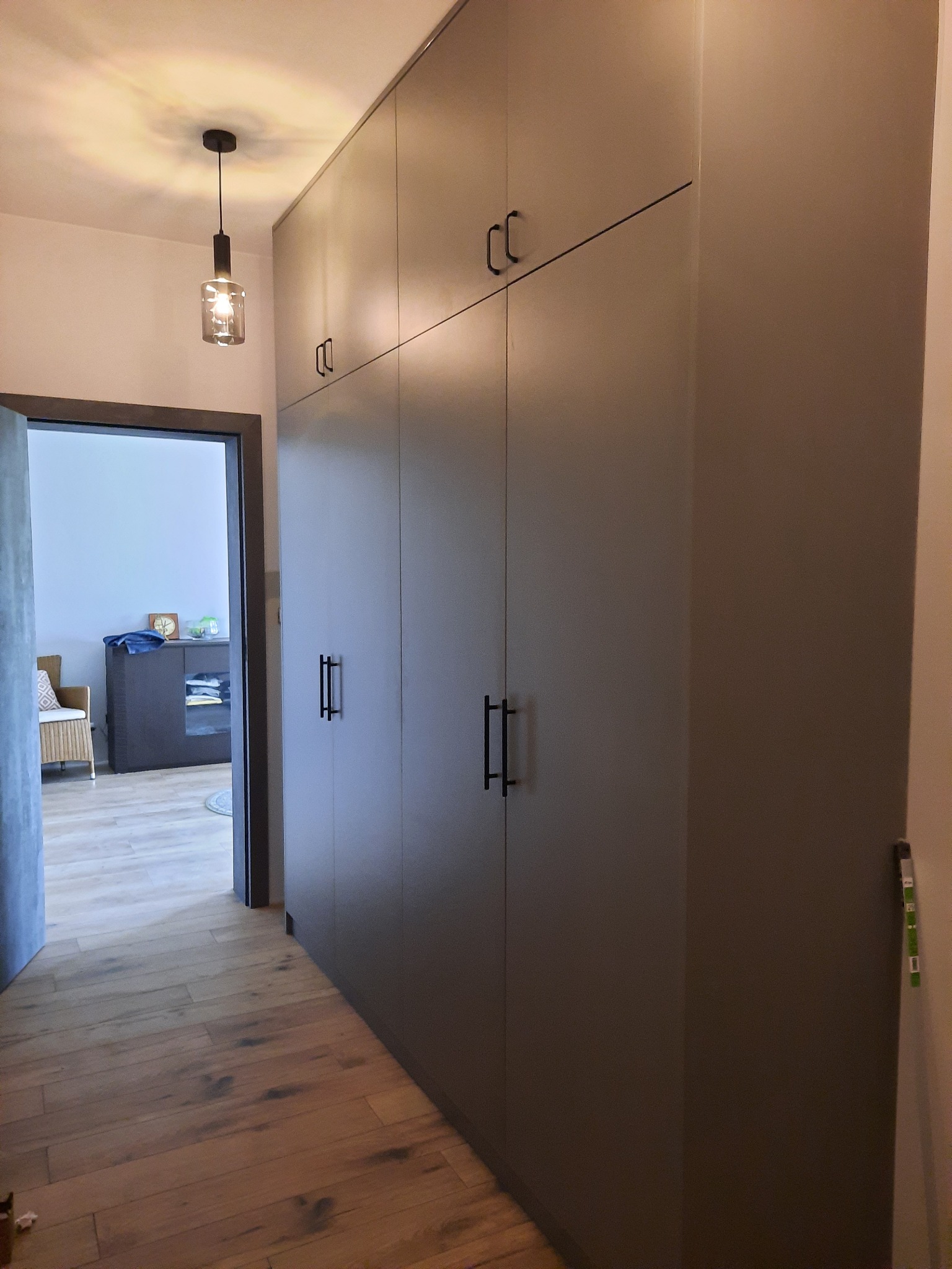 Wysoka, szara szafa wnękowa z minimalistycznymi, czarnymi uchwytami, widoczna na tle drewnianej podłogi i otwartych drzwi do pokoju z meblami.