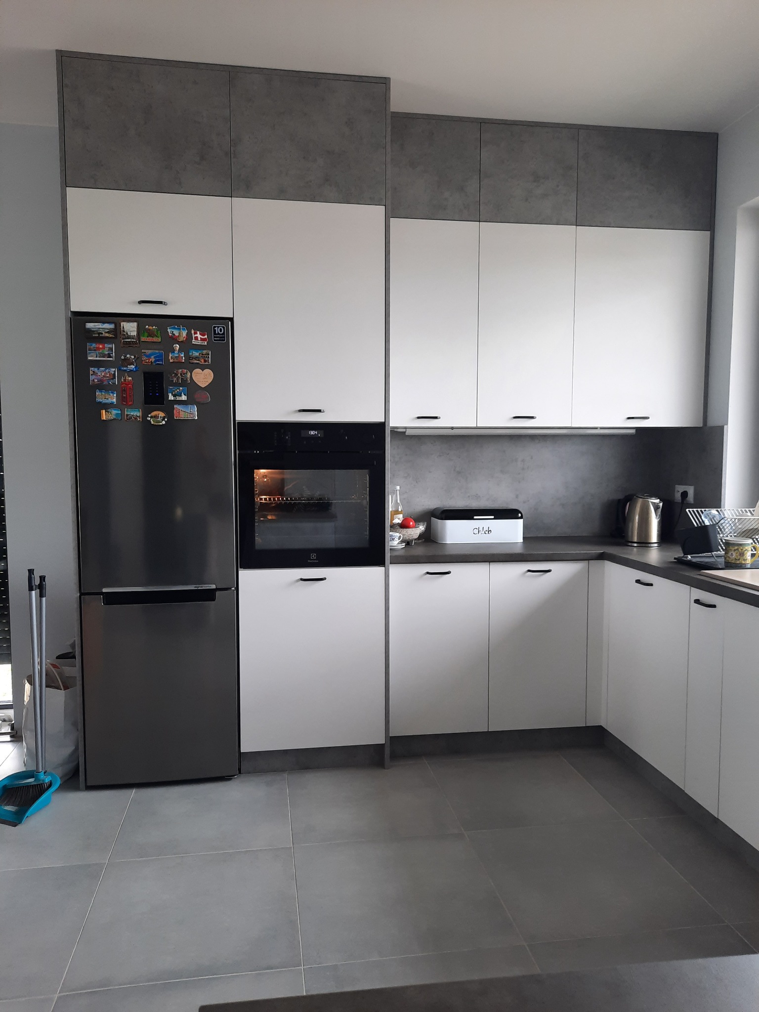 Nowoczesna kuchnia z białymi szafkami i szarym blatem, widoczna lodówka z magnesami i piekarnik z piekącym się ciastem.