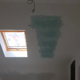 Poddasze w trakcie remontu, widoczne załatane ściany, okno dachowe z zabezpieczeniami i wisząca żarówka.