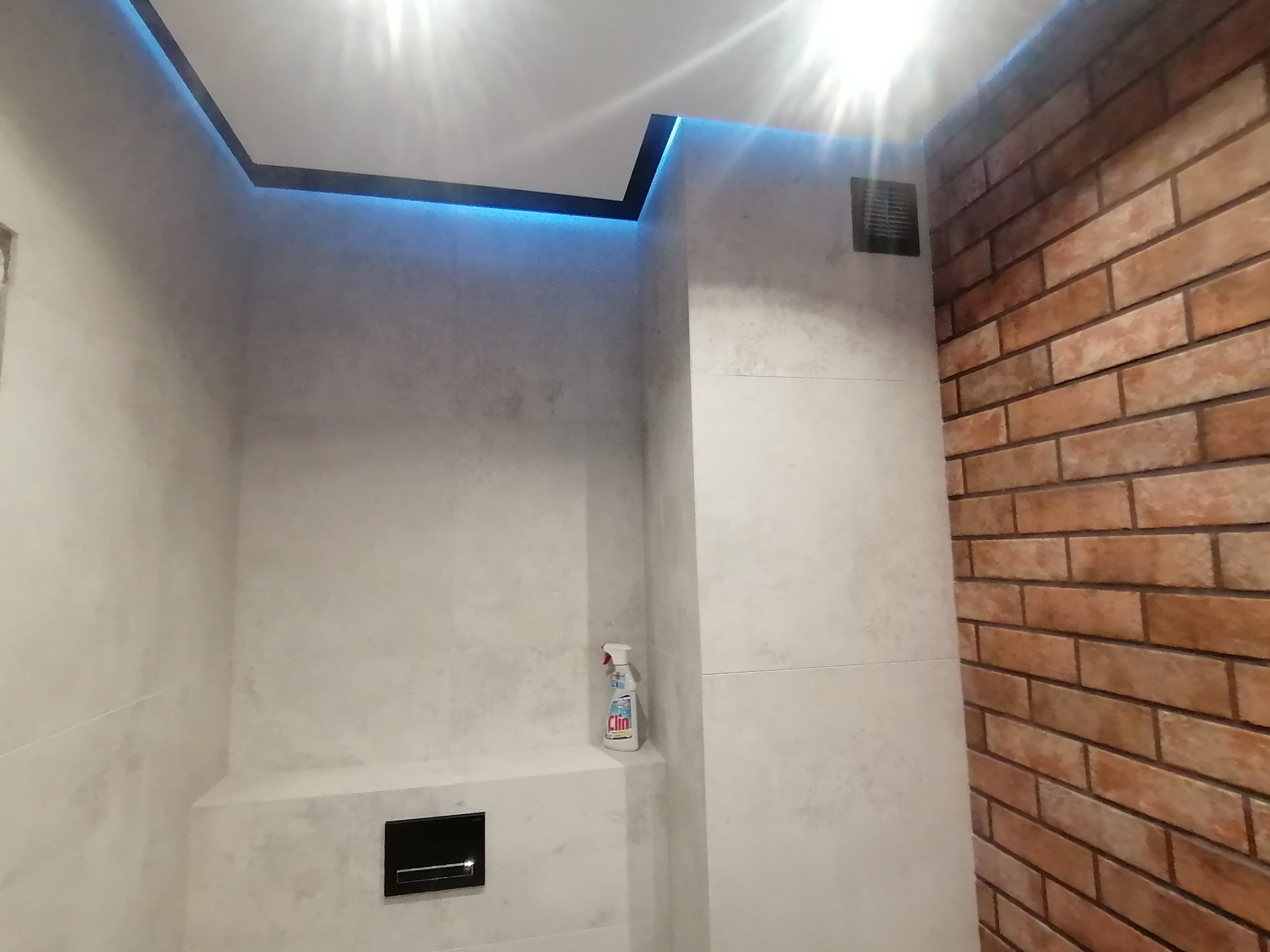 Wykończona łazienka z podświetlanym sufitem, szarą płytką imitującą beton, ceglaną ścianą, czarnym przyciskiem spłuczki i butelką płynu Clin na półce.