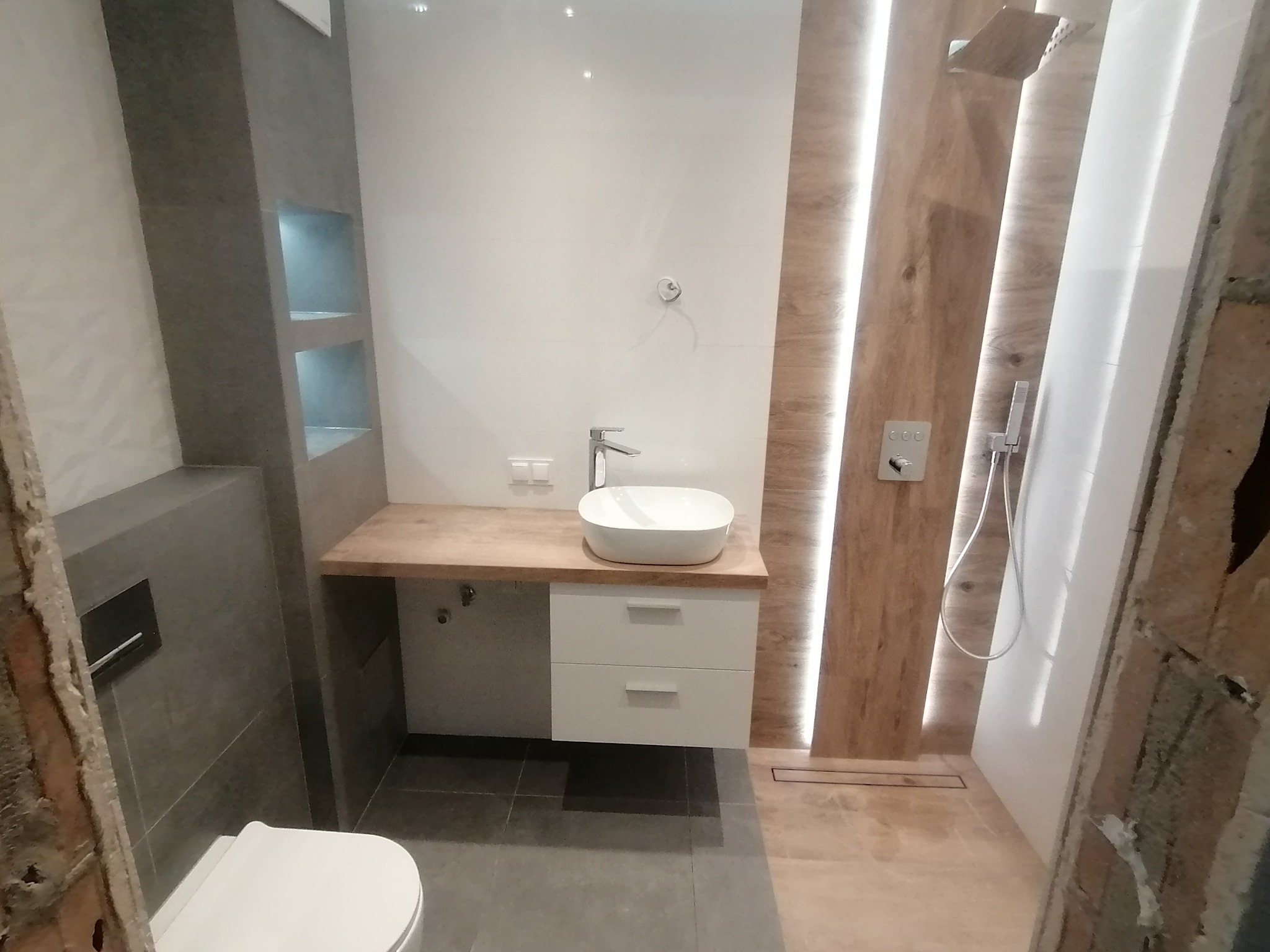 Nowoczesna łazienka w trakcie wykończenia, widoczna umywalka nablatowa na szafce z szufladami, szara zabudowa z półkami oraz kabina prysznicowa z panelami imitującymi drewno.