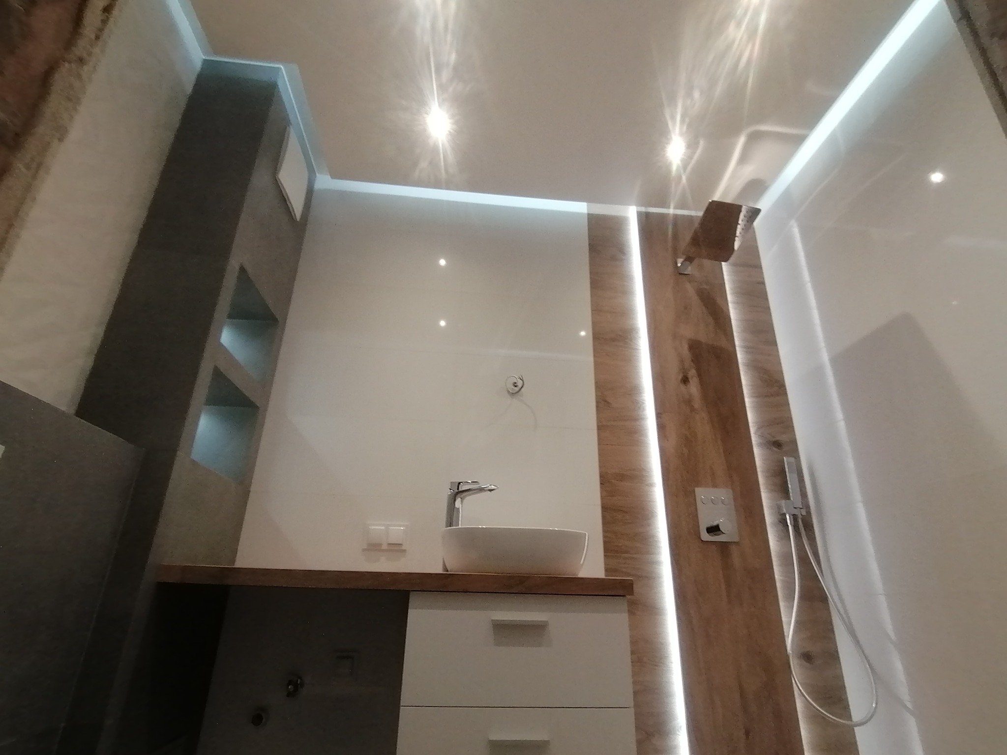 Nowoczesna łazienka z umywalką nablatową, drewnopodobnymi panelami ściennymi z oświetleniem LED oraz kabiną prysznicową typu walk-in.