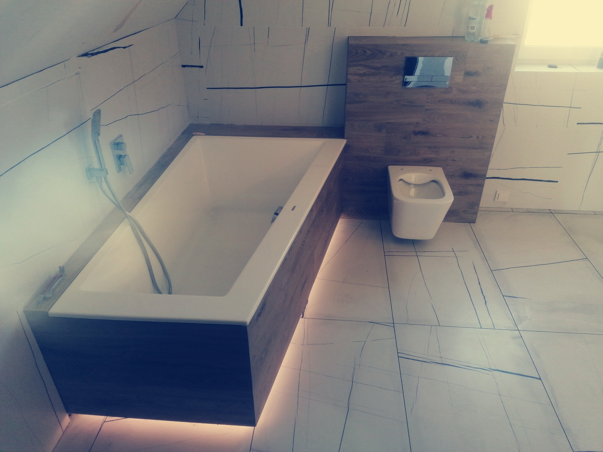 Nowoczesna łazienka z wanną obudowaną drewnem, podświetleniem LED, wiszącą toaletą i biało-szarą glazurą z czarnymi liniami.