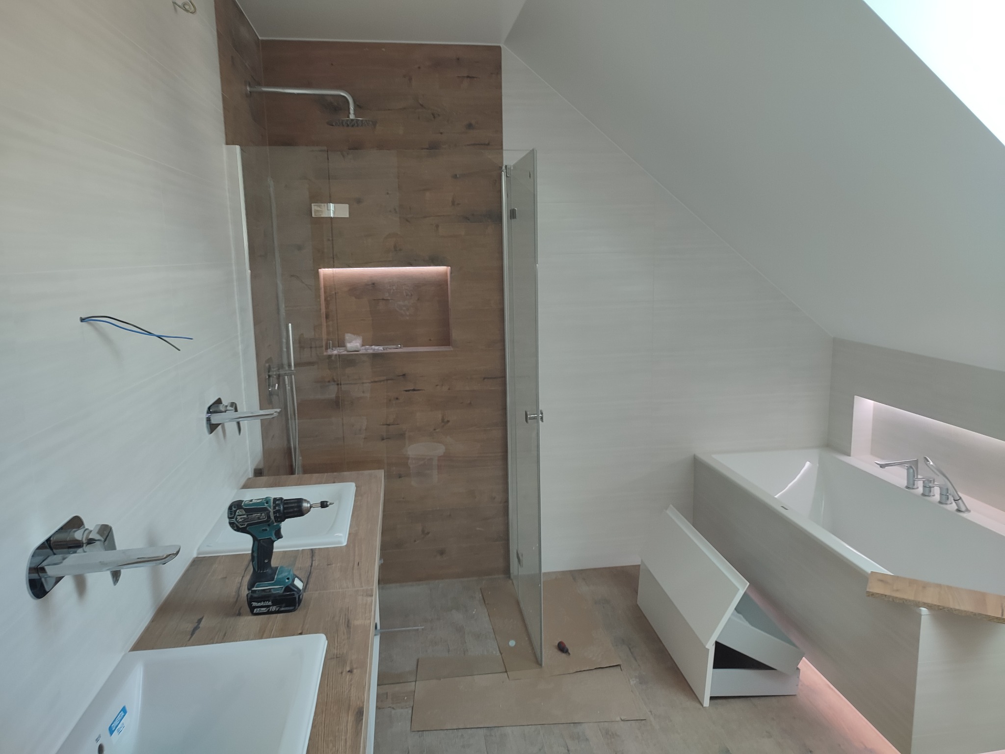 Nowoczesna łazienka w trakcie wykańczania: białe płytki, drewnopodobne akcenty, kabina prysznicowa typu walk-in, umywalki nablatowe z bateriami, wanna z podświetleniem LED, wiertarka na blacie.