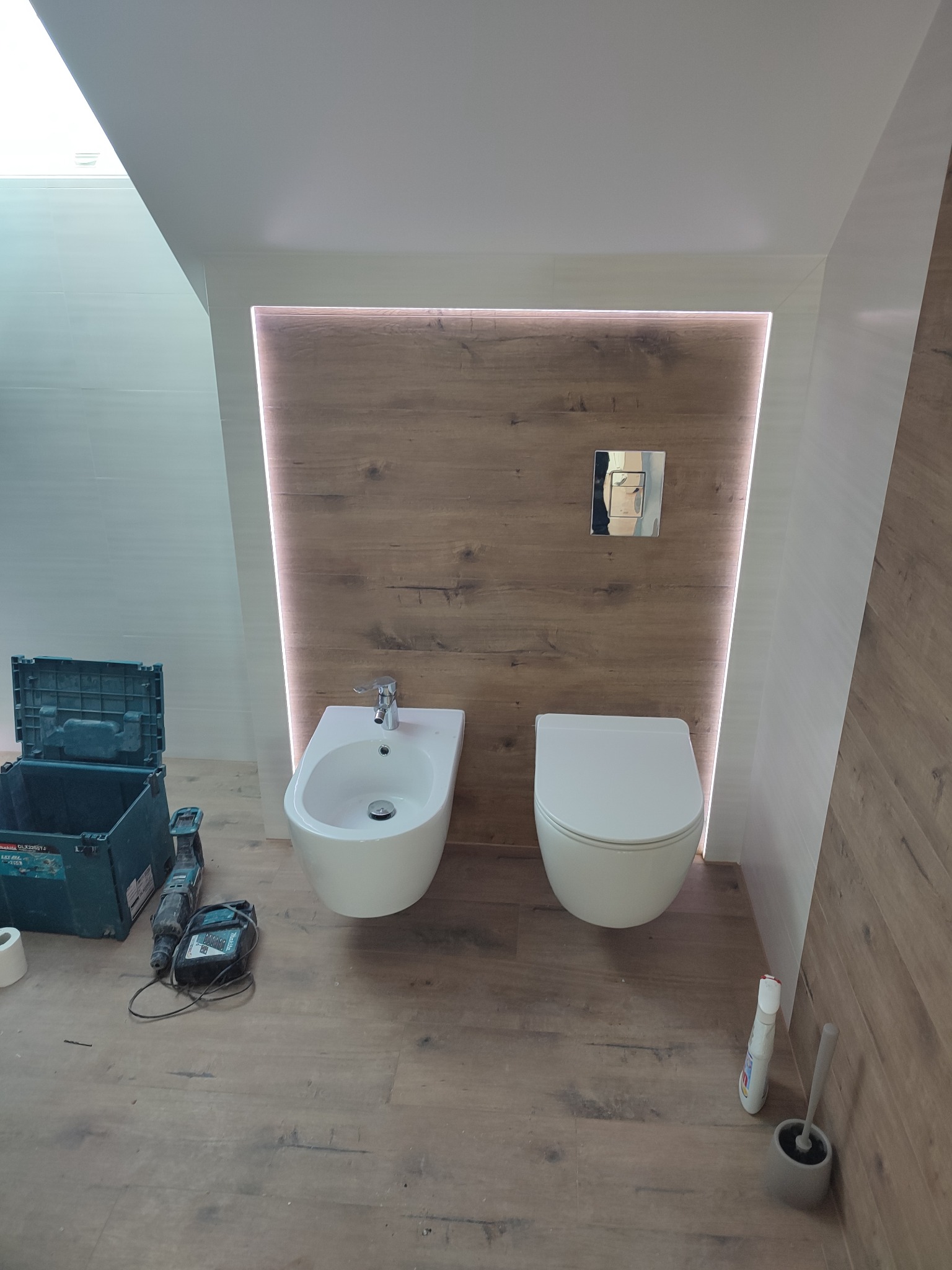 Biały bidet i podwieszana toaleta na tle drewnopodobnej ściany z podświetleniem LED w łazience w Gdyni, obok narzędzia i otwarty kufer narzędziowy.