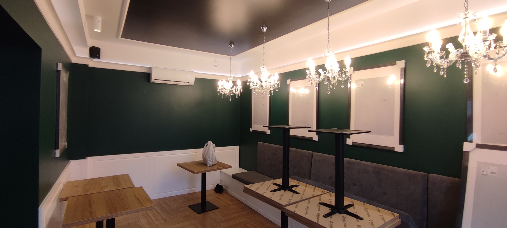 Wnętrze lokalu gastronomicznego z ciemnozielonymi ścianami, białymi boazeriami, drewnianą podłogą i sufitem z czarnym panelem centralnym, oświetlone kryształowymi żyrandolami, z widocznymi...