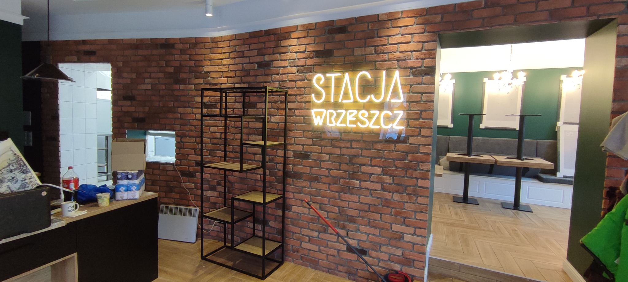 Wnętrze lokalu w trakcie wykańczania z ceglaną ścianą, neonem 'Stacja Wrzeszcz', metalowym regałem i widokiem na salę z stolikami.