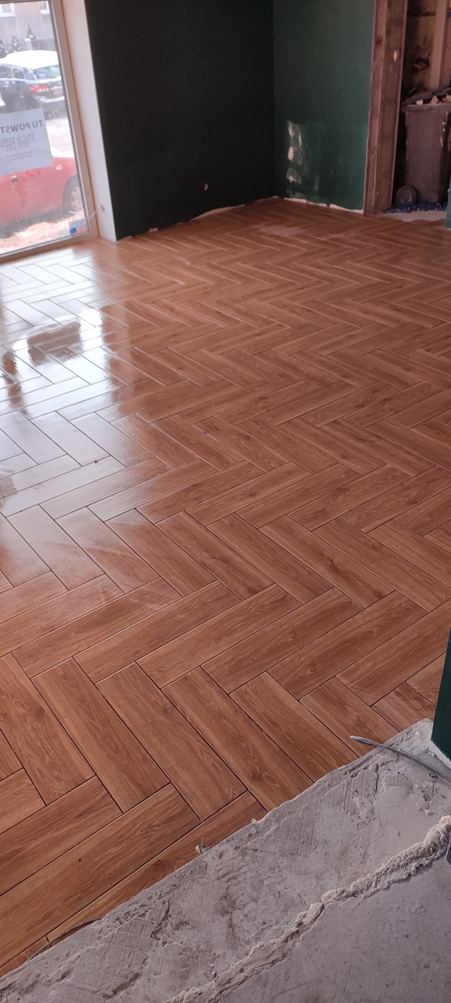 Ułożona parkietem ceramicznym podłoga w jodełkę, widoczna szara, surowa wylewka przy ścianie i fragment ciemnozielonej ściany.