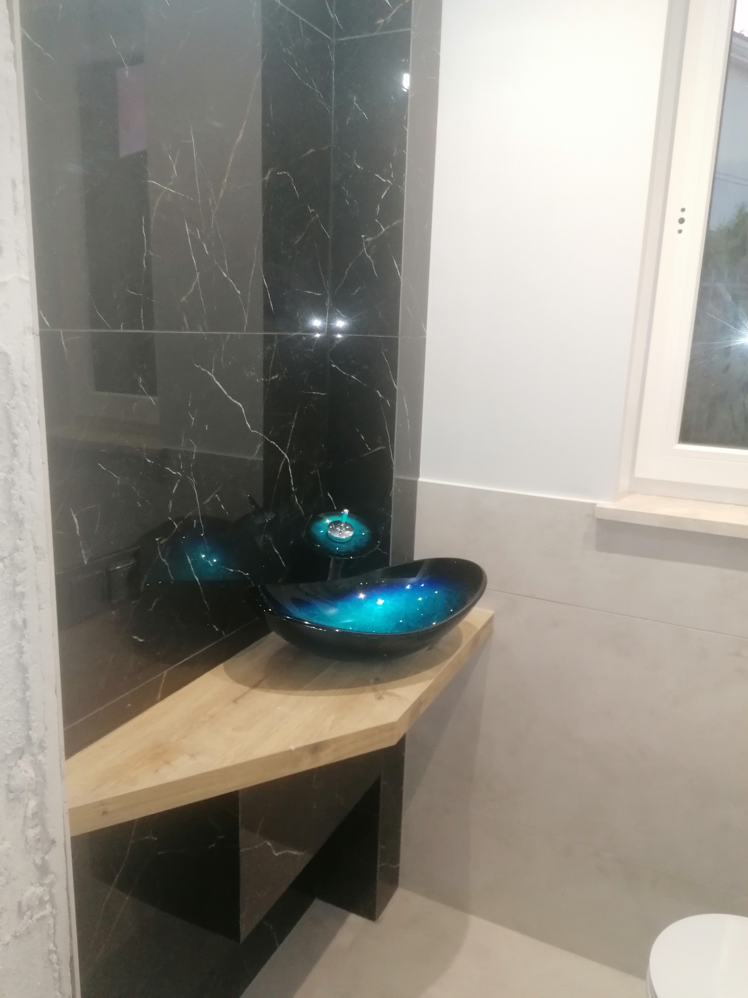 Nowoczesna łazienka z umywalką w kolorze turkusowym, zamontowaną na drewnianym blacie, z czarnymi płytkami imitującymi marmur na ścianie