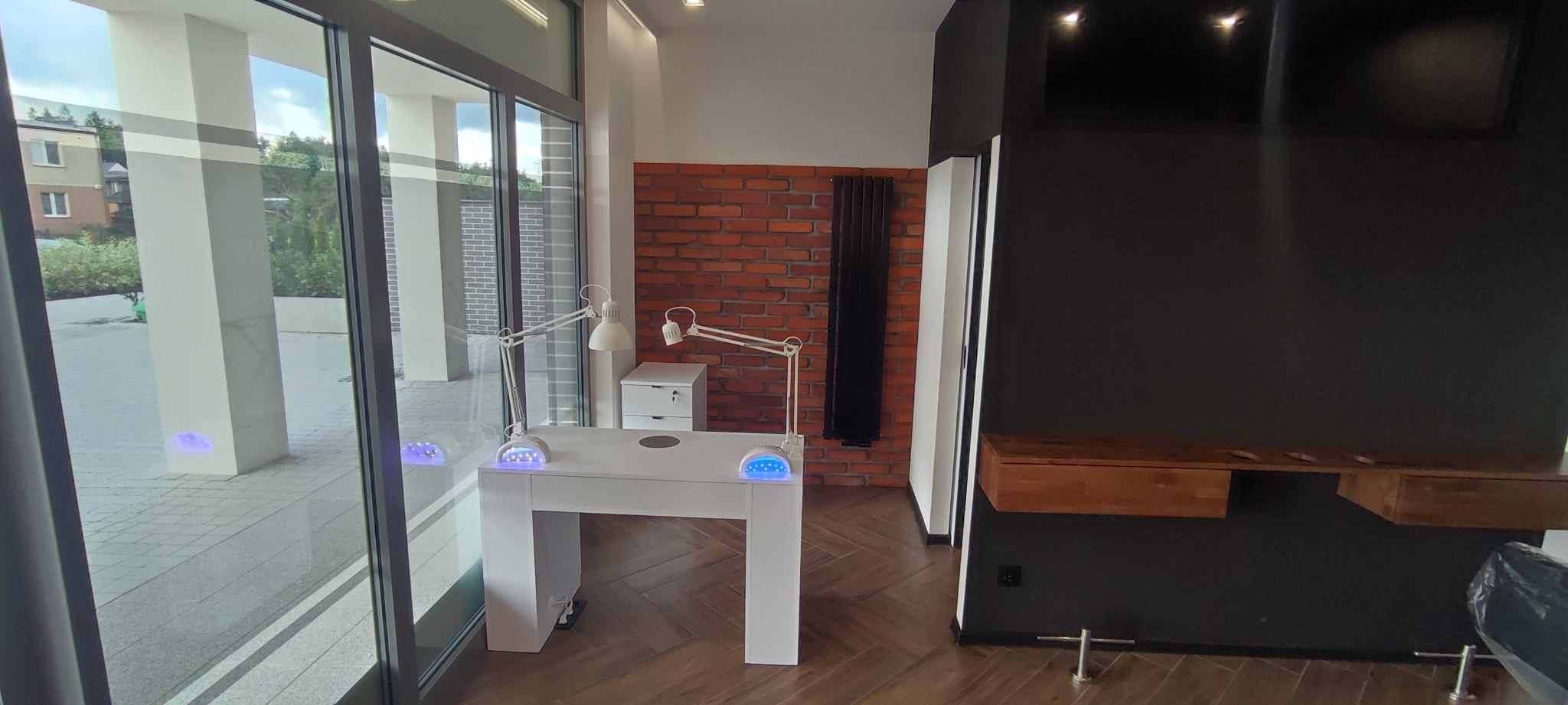 Nowoczesny salon kosmetyczny: białe stanowisko do manicure z lampami UV, ceglana ściana i minimalistyczne meble w odcieniach brązu i czerni, widok przez przeszklone drzwi na zewnątrz.