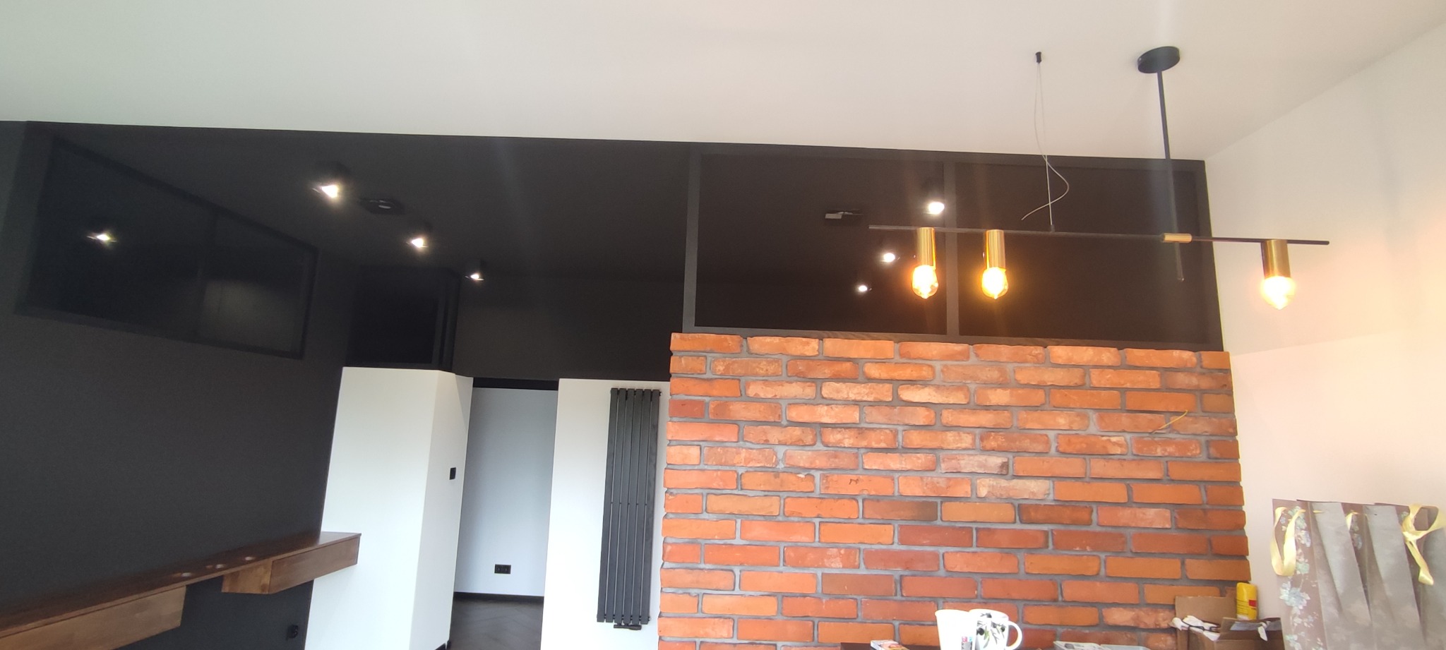 Nowoczesne wnętrze z ceglaną ścianą, czarnym sufitem z oświetleniem punktowym, designerską lampą wiszącą i grzejnikiem panelowym.