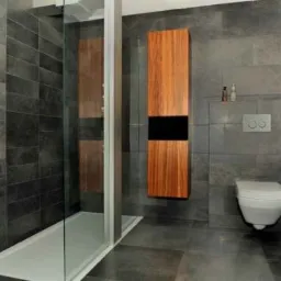 Nowoczesna łazienka z szarymi, prostokątnymi płytkami, kabiną prysznicową ze szklanymi drzwiami, podwieszaną szafką z drewna z czarnym pasem oraz podwieszanym WC.