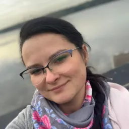 Portret uśmiechniętej kobiety w okularach, z szarym szalem w różowe kwiaty, na tle pochmurnego nieba i wody.