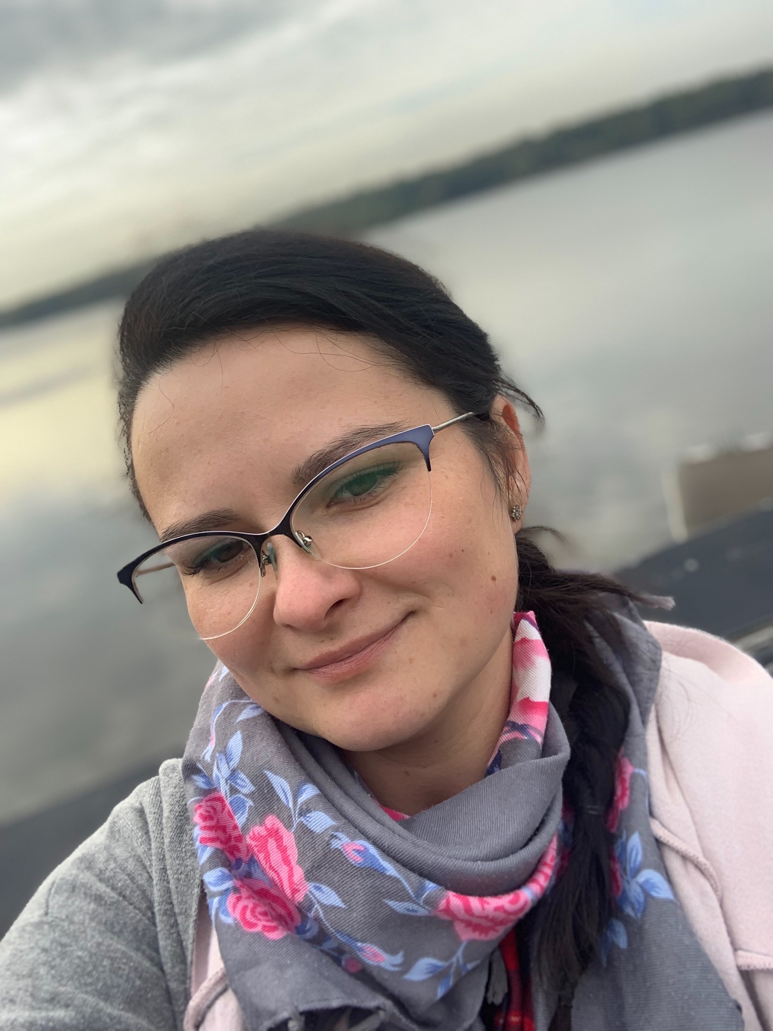 Portret uśmiechniętej kobiety w okularach, z szarym szalem w różowe kwiaty, na tle pochmurnego nieba i wody.