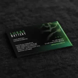 Czarno-zielona wizytówka firmy Lukas Battery leży na ciemnoszarym, teksturowanym tle. Wizytówka zawiera logo, dane kontaktowe i informacje o dystrybucji baterii.