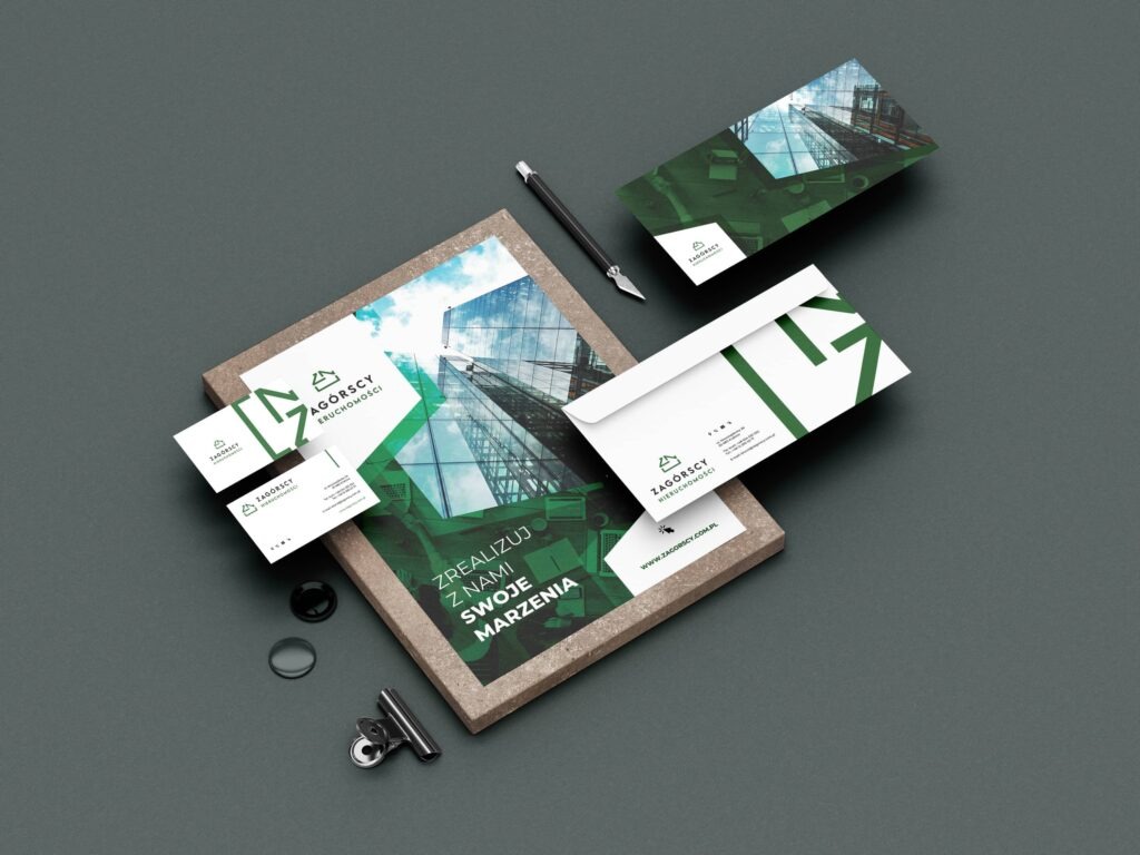 Zestaw materiałów firmowych z logo firmy Zagórscy Nieruchomości: wizytówki, ulotka w ramce, koperta i karta pocztowa, ułożone na szarym tle z dodatkami biurowymi.