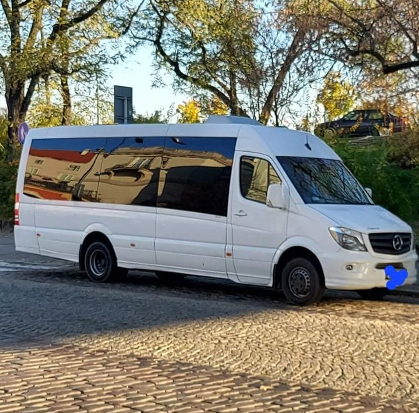 Biały bus z przyciemnianymi szybami, zaparkowany na brukowanej ulicy w słoneczny dzień, z widocznym odbiciem budynku w oknach.