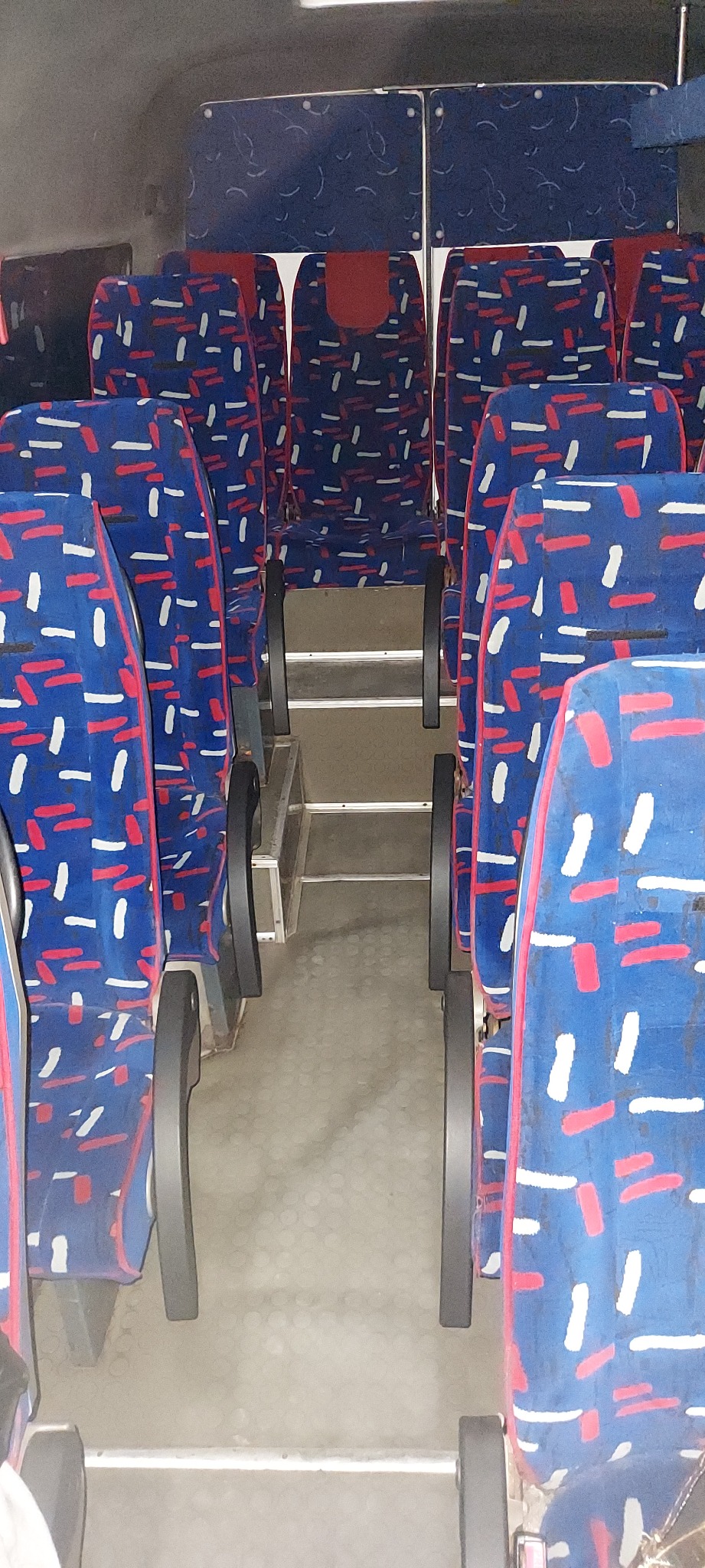 Wnętrze autobusu z niebieskimi siedzeniami w białe i czerwone paski, widok na przejście między rzędami, metalowe stopnie.