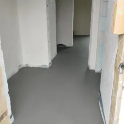 Świeżo wylana, gładka, szara posadzka w remontowanym wnętrzu z widocznymi surowymi ścianami i drewnianą konstrukcją ościeżnicy z boku kadru.