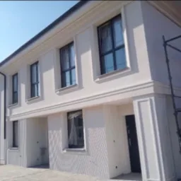 Biała elewacja szeregowca z czarnymi ramami okiennymi, zdobiona sztukaterią, z widocznymi rusztowaniami po prawej stronie.