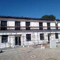 Malowanie elewacji dwukondygnacyjnego budynku z rusztowania, widoczne zabezpieczone okna, farby i narzędzia malarskie na ziemi, dwóch pracowników w trakcie pracy.