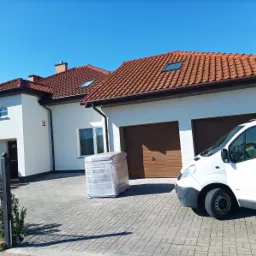 Biały, parterowy dom z czerwoną dachówką i brązowymi bramami garażowymi; na podjeździe zapakowany materiał budowlany i biały bus z otwartymi drzwiami.