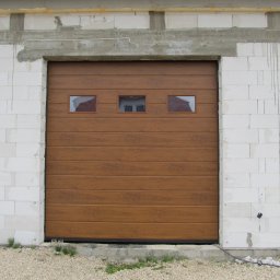 Phtu Control - Brązowa segmentowa brama garażowa z trzema małymi okienkami, zamontowana w surowym murze z betonu komórkowego, widoczne ślady zaprawy.