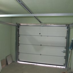 Phtu Control - Montaż segmentowej bramy garażowej: widoczne prowadnice, brama częściowo otwarta, sufit i ściany w kolorze pistacjowym, narzędzia na parapecie okna.