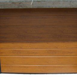 Phtu Control - Brązowa brama garażowa segmentowa z widoczną fakturą drewna, zamontowana w surowym otworze budynku z betonu komórkowego.