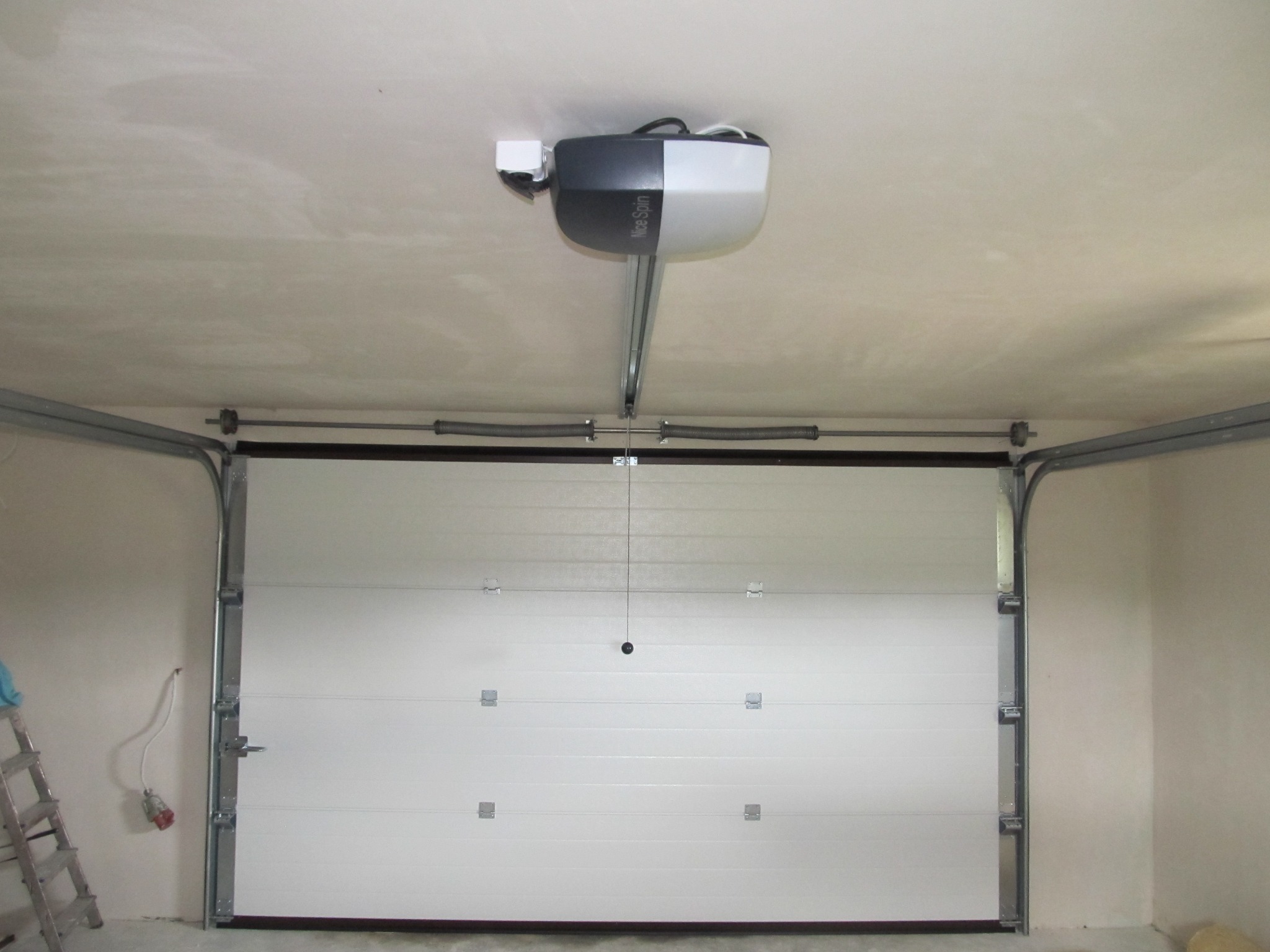 Zamknięta biała brama garażowa segmentowa z napędem Nice Spin zamontowanym pod sufitem garażu, widoczna drabina aluminiowa z boku.