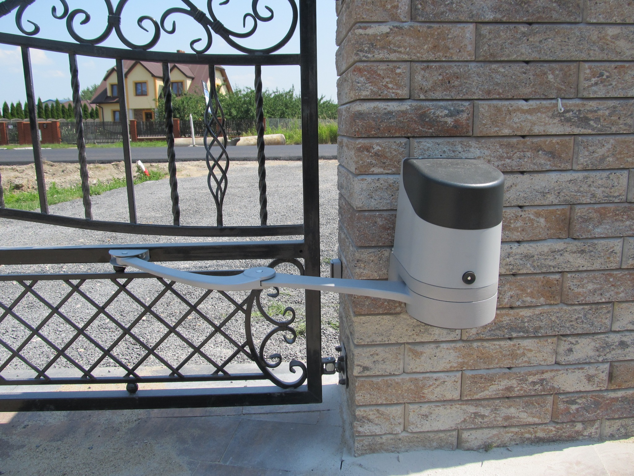 Kute, czarne skrzydło bramy wjazdowej z automatycznym napędem, zamontowanym na murowanym słupku z cegły piaskowej, na tle domu jednorodzinnego.