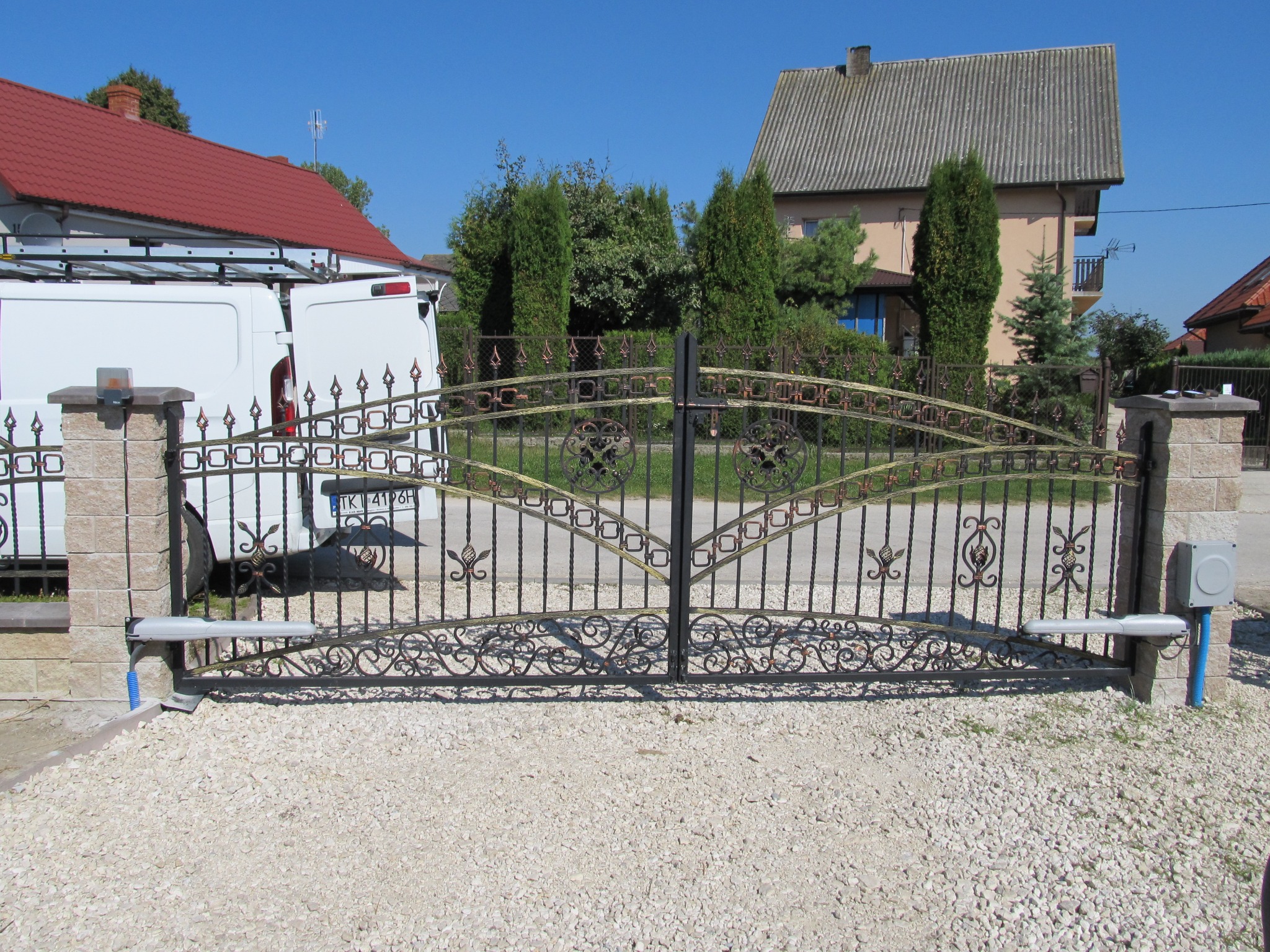 Ozdobna, kuta brama wjazdowa z automatyką, zamontowana na posesji z jasną nawierzchnią żwirową, w tle budynki mieszkalne i biały samochód dostawczy z otwartymi drzwiami.