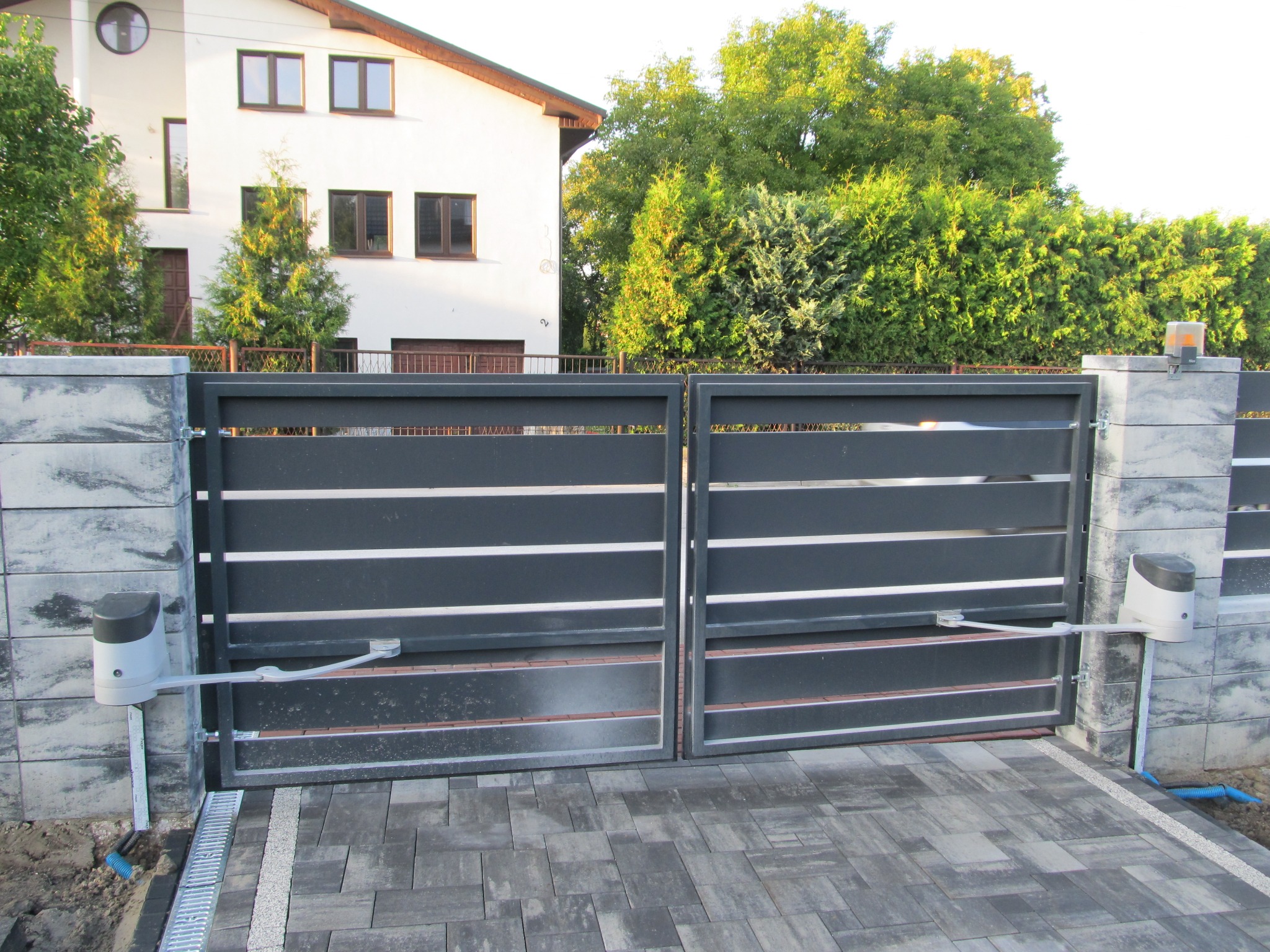 Nowoczesna, dwuskrzydłowa brama wjazdowa z automatyką, szara, poziome panele, kostka brukowa na podjeździe, widoczne siłowniki bramy zamontowane na słupkach.