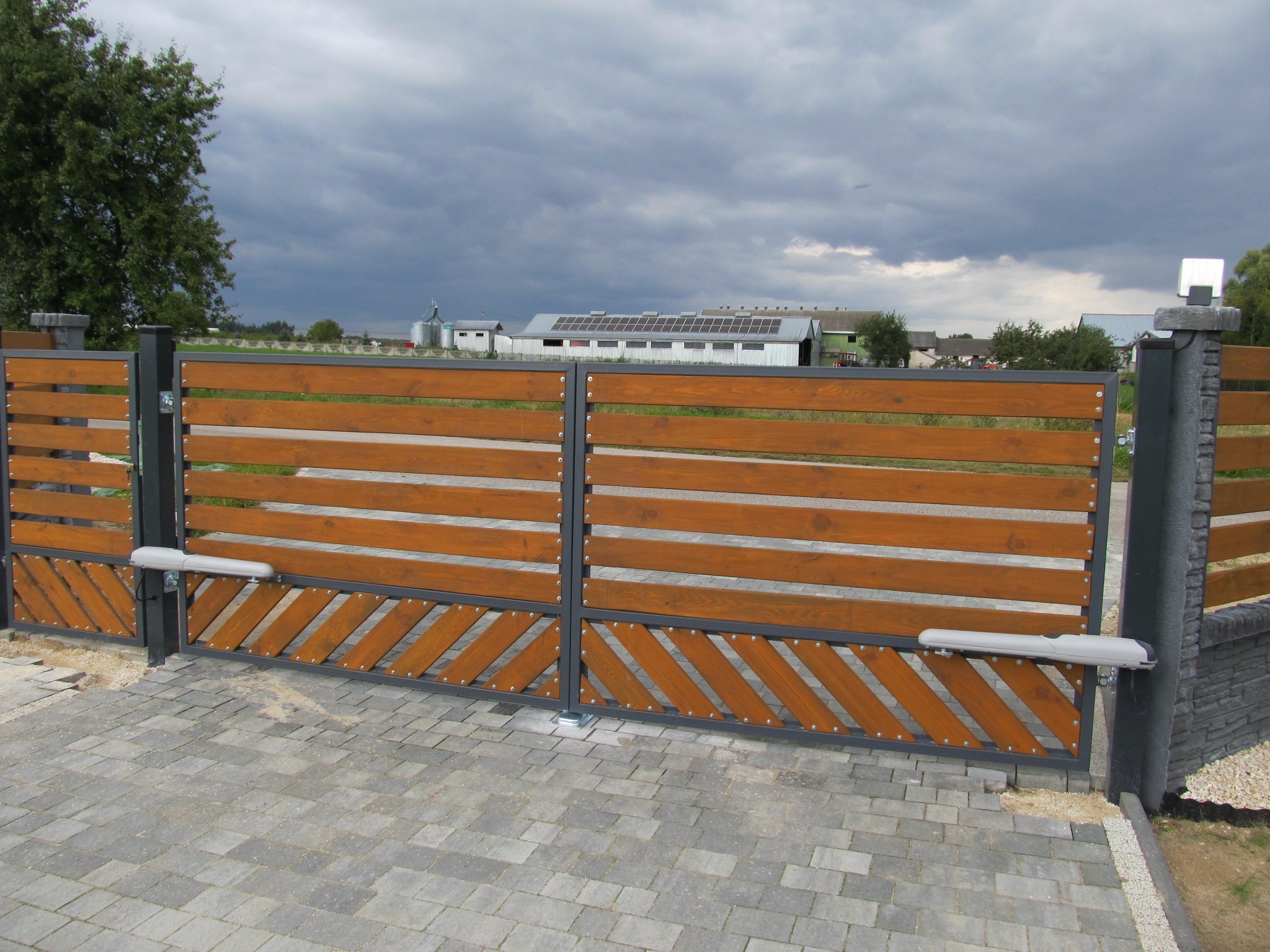Dwuskrzydłowa brama wjazdowa z automatyką, drewniane wypełnienie z poziomych desek i ukośnych elementów w dolnej części, szara kostka brukowa na podjeździe, pochmurne niebo w tle.
