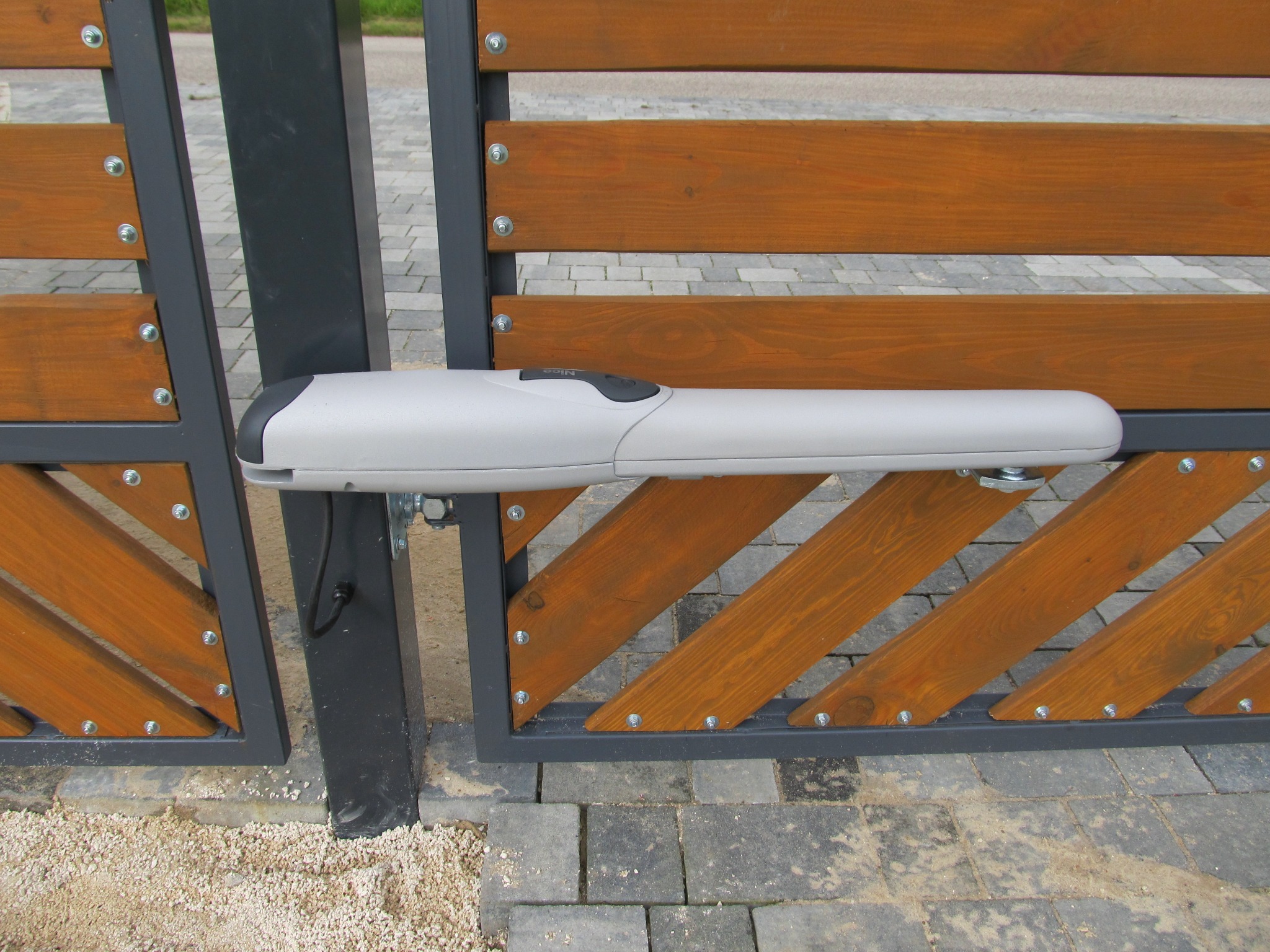 Szary automat do bramy skrzydłowej zamontowany na drewnianej bramie z metalową ramą, widoczne śruby mocujące.