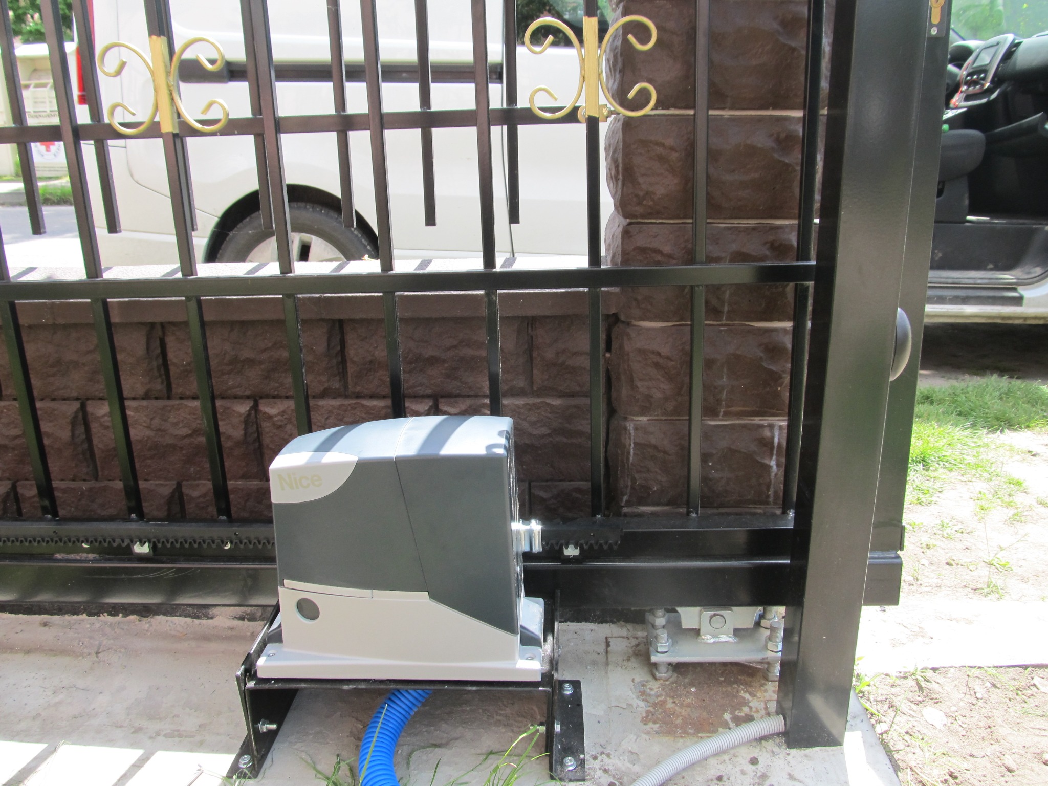 Zamontowany napęd Nice do bramy przesuwnej z widocznym mechanizmem zębatym i okablowaniem, na tle ogrodzenia z cegły i pojazdu.